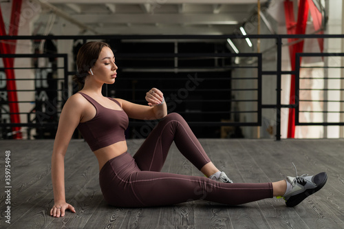A sporty woman is resting sitting on the floor and chatting on a smartphone. Girl listens to music on a wireless headphone during a fitness workout. Mobile application with radio.