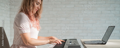 A focused woman plays the electronic piano and watches an online course on her laptop. Distance learning music quarantined. Stay home. A lesson in playing a keyboard instrument.