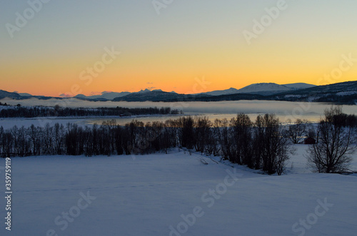 ice fog over calm fjord with strong colorful sunset sky and snowy mountain range in winter © Arcticphotoworks