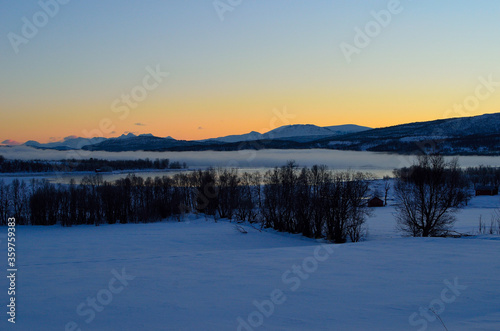 ice fog over calm fjord with strong colorful sunset sky and snowy mountain range in winter