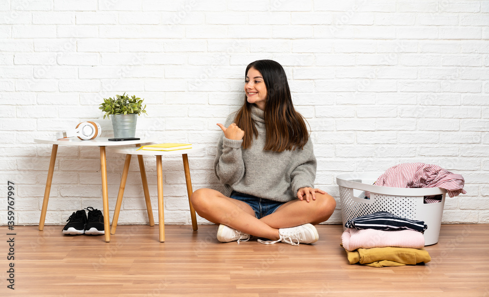 Young woman sitting on the floor at indoors with clothes basket pointing to the side to present a product Young woman sitting on the floor at indoors with clothes basket pointing to the side to present a product