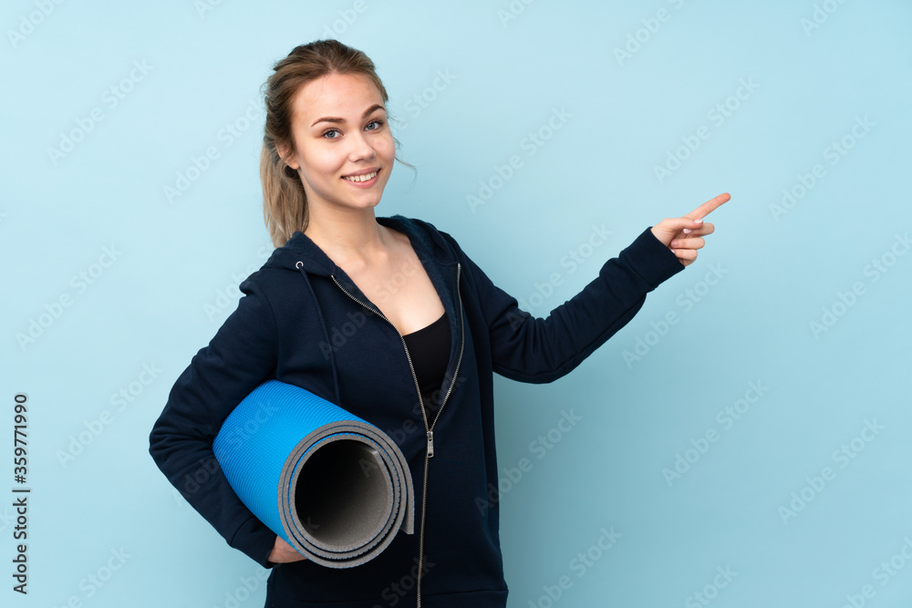 Teenager Russian girl holding mat isolated on blue background pointing finger to the side and presenting a product Teenager Russian girl holding mat isolated on blue background pointing finger to the side and presenting a product