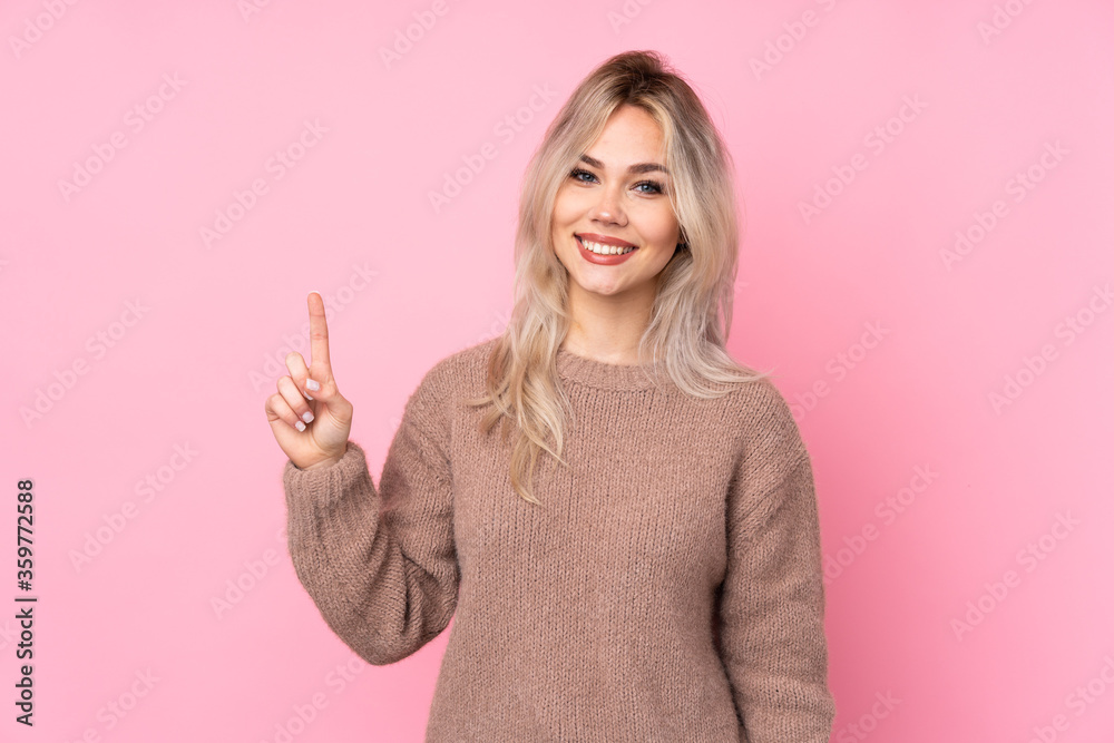 Teenager blonde girl wearing a sweater over isolated pink background showing and lifting a finger in sign of the best