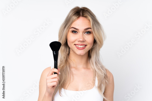 Teenager blonde girl over isolated white background holding makeup brush and whit happy expression
