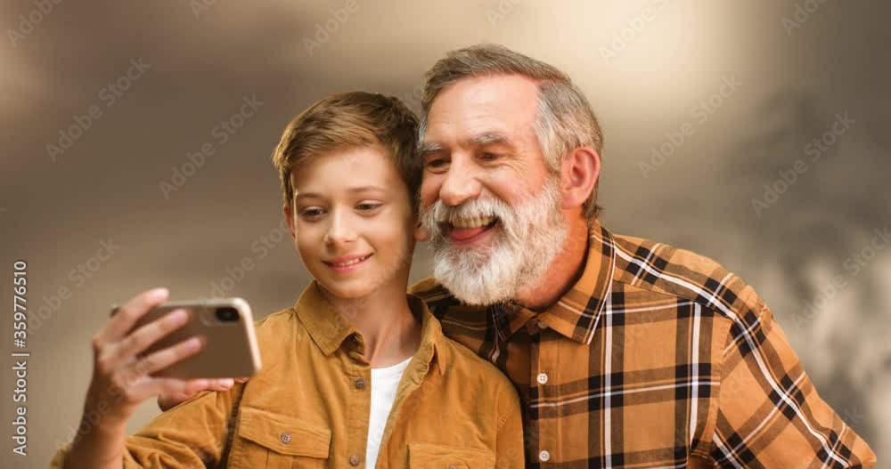 Caucasian teen happy boy taking selfie photo on smartphone camera together with senior grandfather with gray hair and beard. Grandpa and grandson posing to phone and mocking faces. Making funny photos Caucasian teen happy boy taking selfie photo on smartphone camera together with senior grandfather with gray hair and beard. Grandpa and grandson posing to phone and mocking faces. Making funny photos