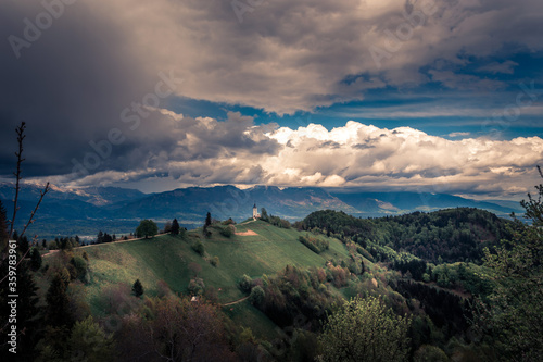Famous Jamnik church on hilltop in summer season with incoming storm clouds. Picturesque landmark in Slovenia. In background Alps mountain range. Green landscape