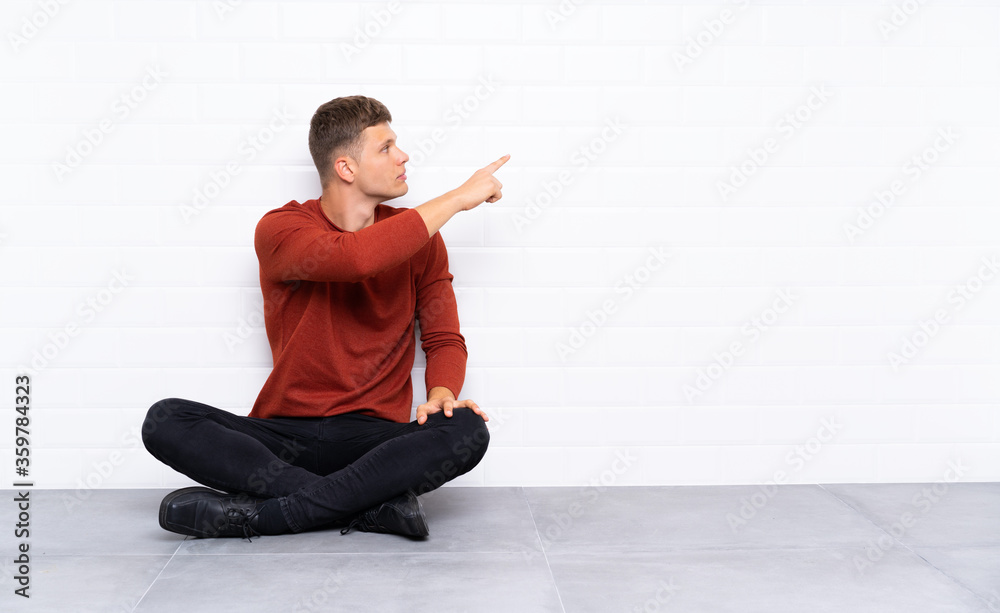 Young handsome man sitting on the floor pointing back with the index finger Young handsome man sitting on the floor pointing back with the index finger