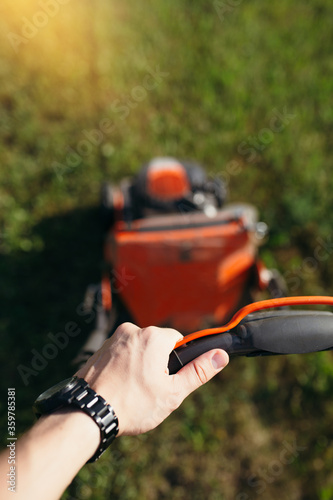 First-person photo. A man's hand holds a lawn mower and mows a lawn outside the city on its plot at sunset.