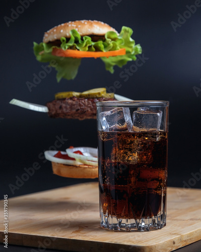 Classic Burger. Still life of a Burger and coke. Drink in a tall glass with ice. The components of the Burger are flying in the air on a black background. Meat cutlet, onion, tomato, lettuce and chees