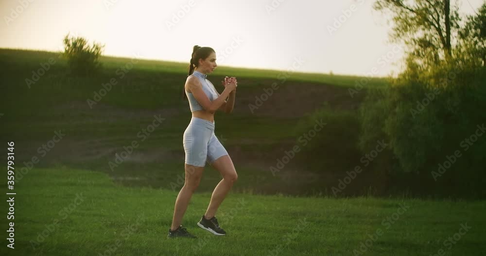 A woman walks on the grass in a Park performing alternate lunges on each leg in motion at sunset in summer A woman walks on the grass in a Park performing alternate lunges on each leg in motion at sunset in summer