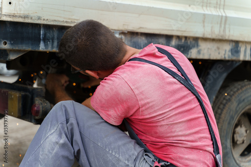 Caucasian man unknown person male adult fixing the back of the truck or vehicle solving problems while sitting under the chassis in day - Mechanic worker repairing truck - back view