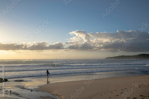 surfer walking along beach with surfboard and sun setting in background
