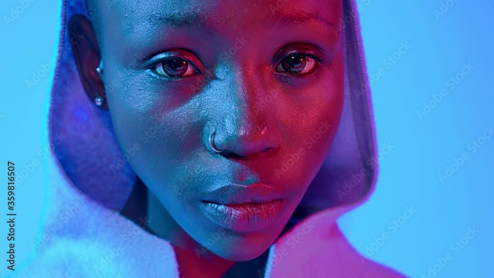 Extreme close-up portrait of a sad young Black woman wearing a hood under blue studio lights.
