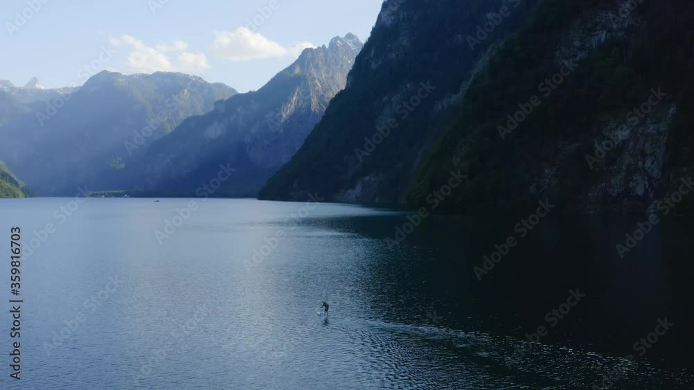 Aerial view Trucking shot, Man paddle boarding SUP on the lake, Scenic view mountain range blue bright sky in the background. Aerial view Trucking shot, Man paddle boarding SUP on the lake, Scenic view mountain range blue bright sky in the background.