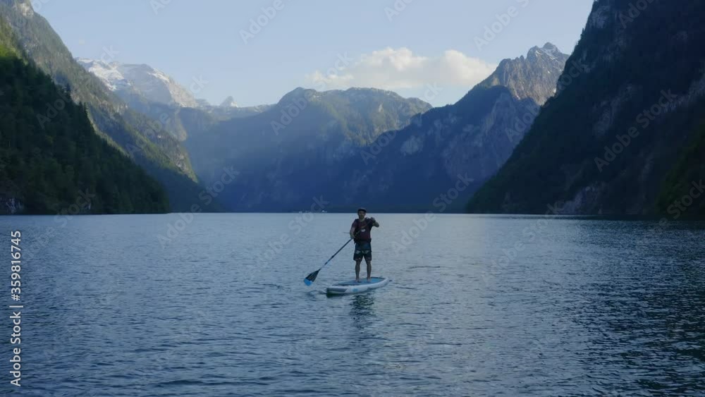Aerial view Moving away shot, a man takes a picture of another man on a paddle board, Scenic view of the lake and mountains in the background. Aerial view Moving away shot, a man takes a picture of another man on a paddle board, Scenic view of the lake and mountains in the background.