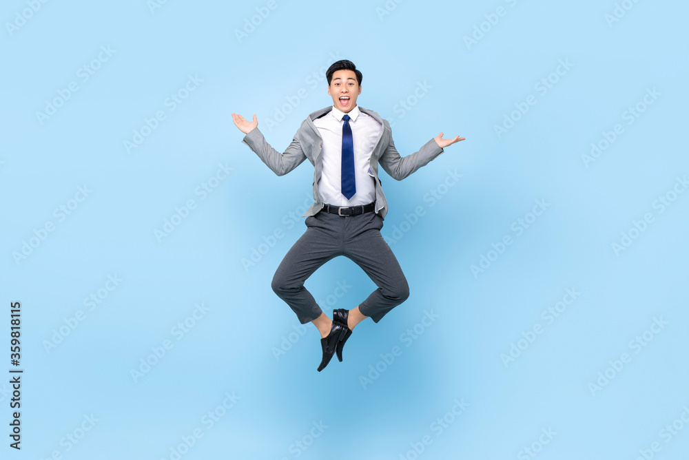 Full length playful portrait of happy ecstatic young Asian businessman jumping in mid-air doing wacky fun gesture in isolated studio blue background