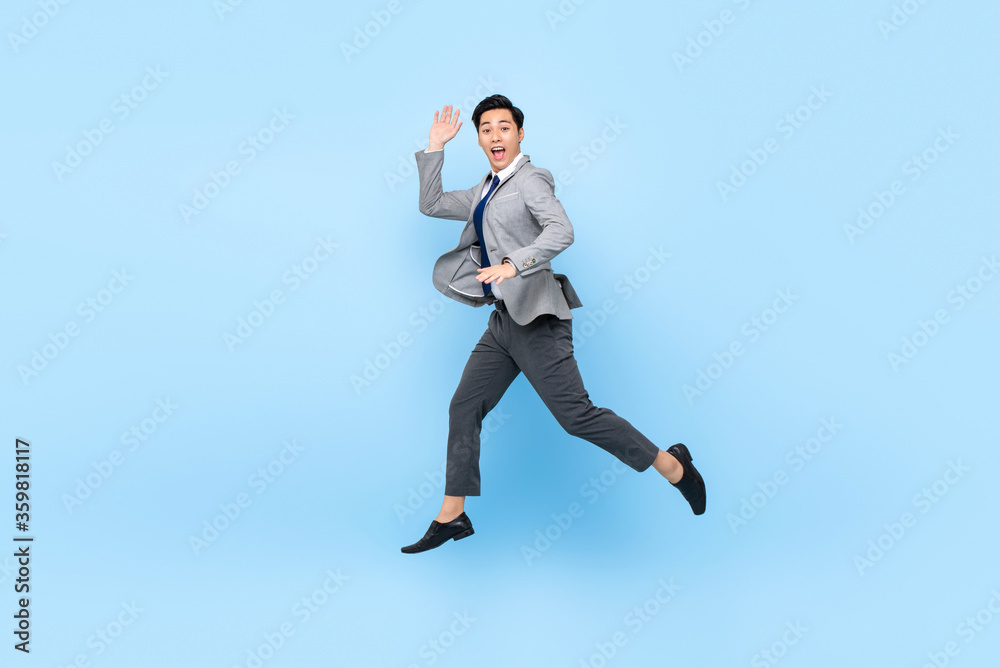 Full length fun portrait of happy ecstatic young Asian businessman jumping in mid-air isolated on studio blue background