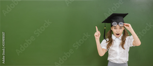 Surpraise young girl wearing graduation hat points away on empty green chalkboard. Empty space for text photo