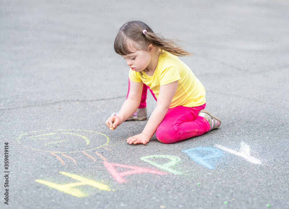 Fototapeta premium Little girl with syndrome down draws with chalk on the asphalt on summer day