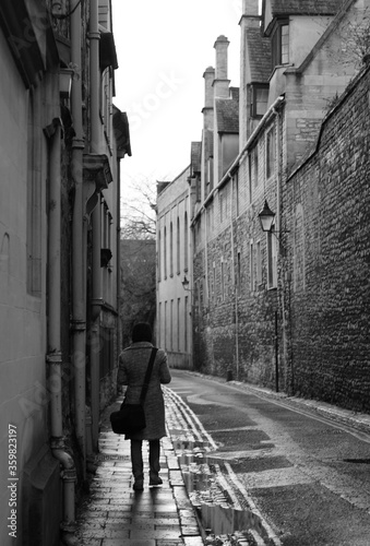 Fototapeta Naklejka Na Ścianę i Meble -  Black and white image showing a woman wearing winter coat and carrying a shoulder bag is walking through a narrow alley with historical buildings on both sides. It is a rainy day with water puddles.