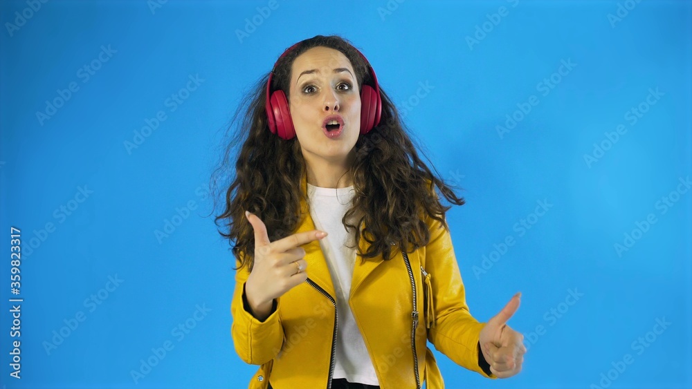 Naklejka premium Young woman in yellow jacket dancing while listening music on headphones in Studio with blue Background.