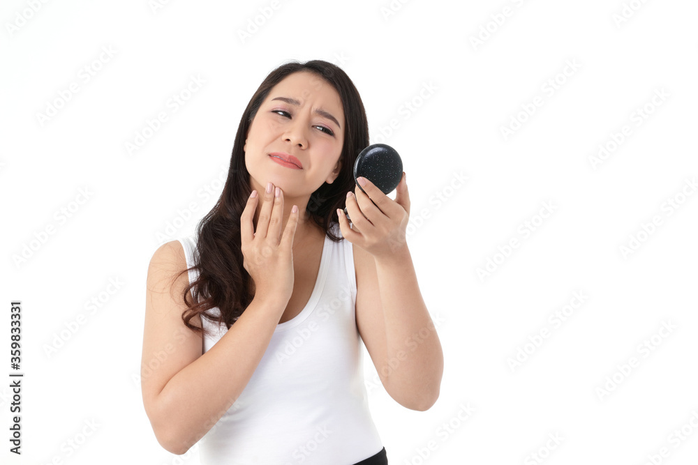A beautiful Asian woman feels stressed. She is holding a mirror, looking at acne on her cheeks. Beauty concept, white background A beautiful Asian woman feels stressed. She is holding a mirror, looking at acne on her cheeks. Beauty concept, white background