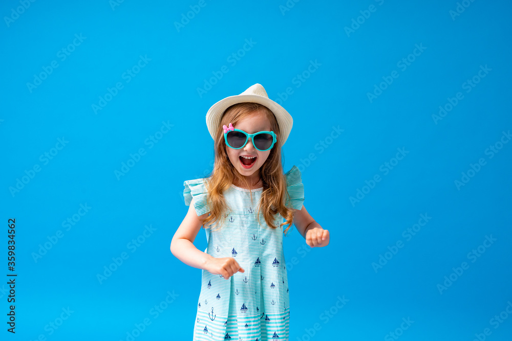 cute little girl in a dress, hat and sunglasses poses on a blue background