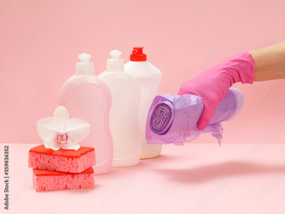 Bottles of dishwashing liquid, sponges and hand with garbage bags on a pink background.
