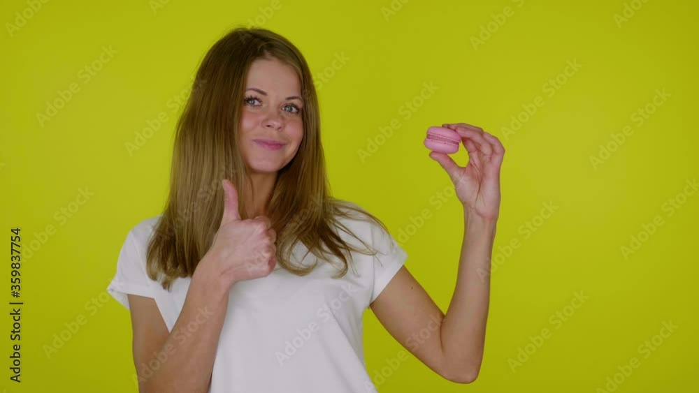 Positive woman in a white T-shirt raises hand with a pink macaroon, shows like and smiles on a yellow background with copy space. Place for text or product. 4K slow motion footage Positive woman in a white T-shirt raises hand with a pink macaroon, shows like and smiles on a yellow background with copy space. Place for text or product. 4K slow motion footage