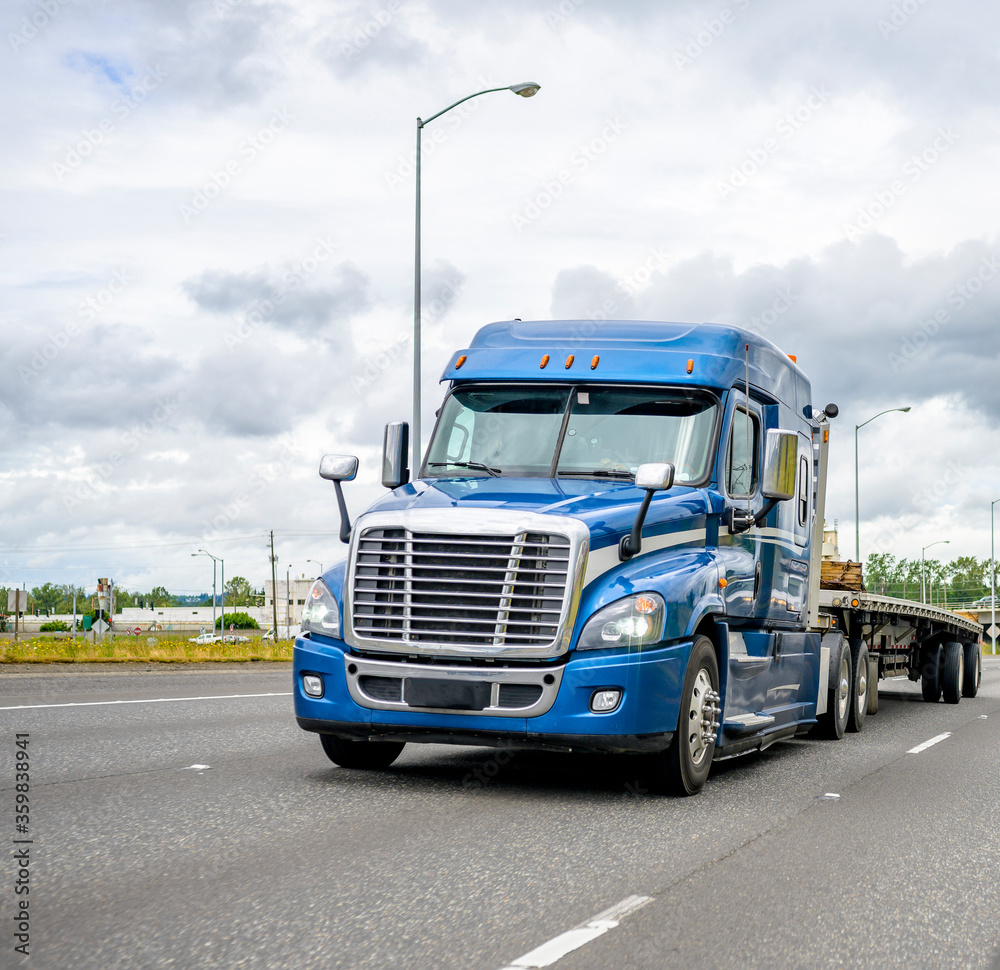 Blue big rig semi truck with flat bed semi trailer running on wide road to warehouse for pick up load for next delivery Blue big rig semi truck with flat bed semi trailer running on wide road to warehouse for pick up load for next delivery