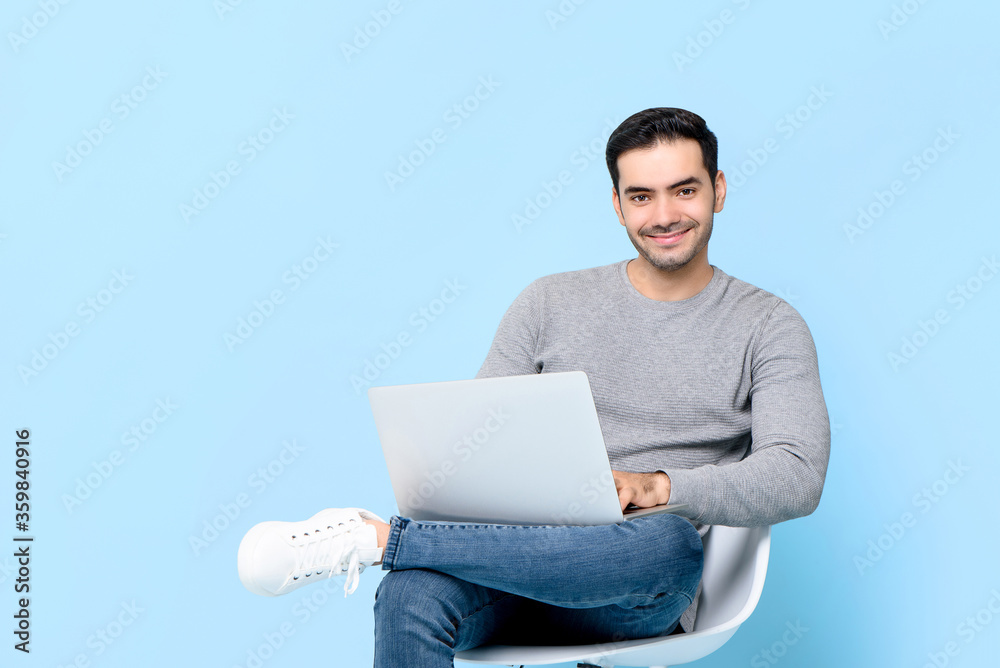 Portrait of smiling young handsome Caucasian man looking at camera while sitting using laptop in isolated studio blue background Portrait of smiling young handsome Caucasian man looking at camera while sitting using laptop in isolated studio blue background