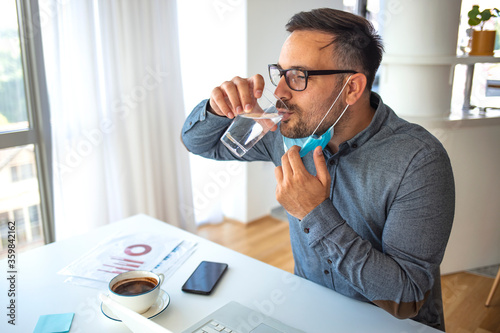 Businessman with mask drinking water at work. Staying hydrated on his business. Businessman having a glass of water while working in office and looking away - dehydration