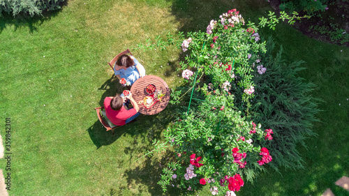 Young couple enjoying food and wine in beautiful roses garden on romantic date, aerial top view from above of man and woman eating and drinking together outdoors in park 