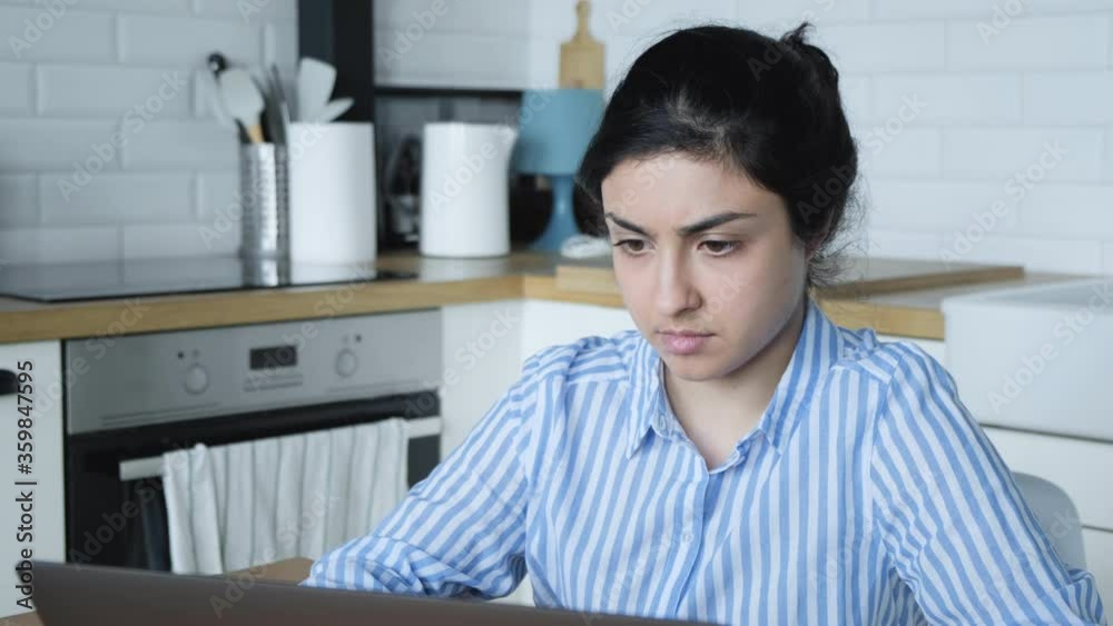 A serious Young Indian girl in a blue striped shirt is working on a computer at home sitting in a bright kitchen