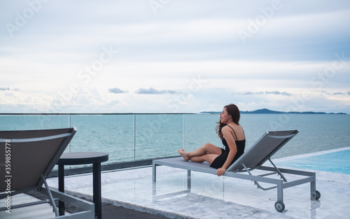 A beautiful young asian woman sitting next to the rooftop pool , looking at the sea and blue sky view