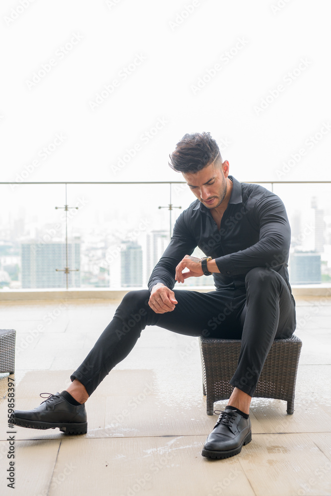 Full body shot of young bearded Persian businessman checking the time while sitting Full body shot of young bearded Persian businessman checking the time while sitting