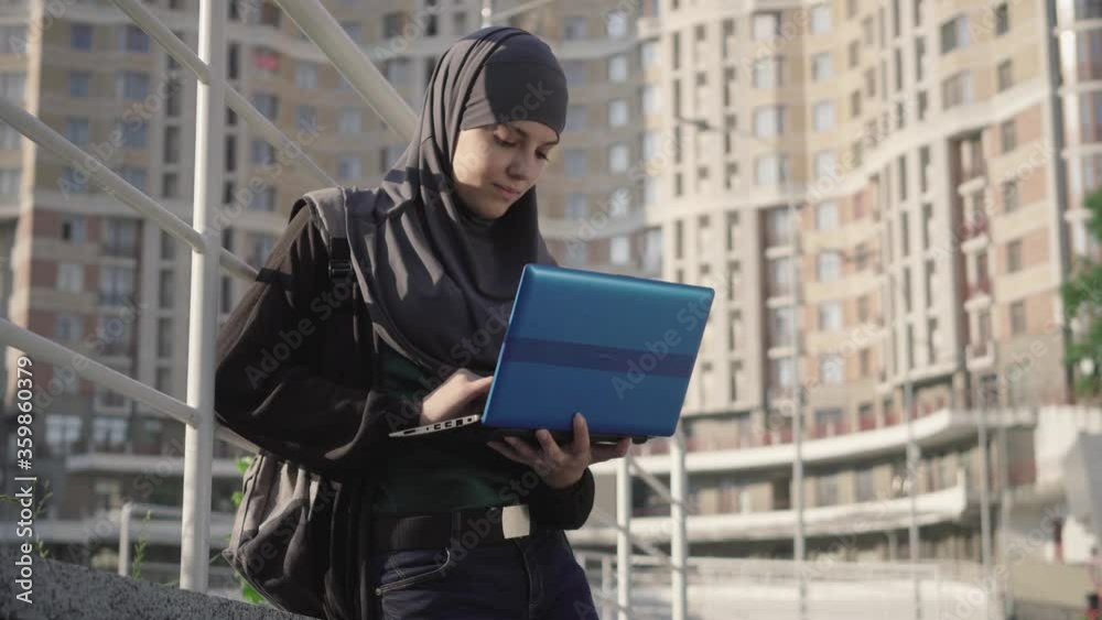Smiling muslim woman in traditional hijab typing on laptop keyboard ...