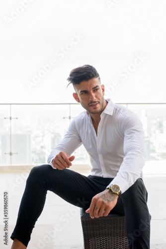 Happy young handsome bearded Persian businessman sitting against view the city