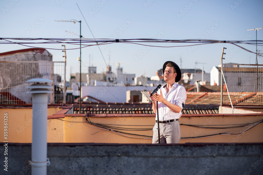 Chico joven con sombrero cantando en la azotea de un edificio con vistas de la ciudad al fondo
