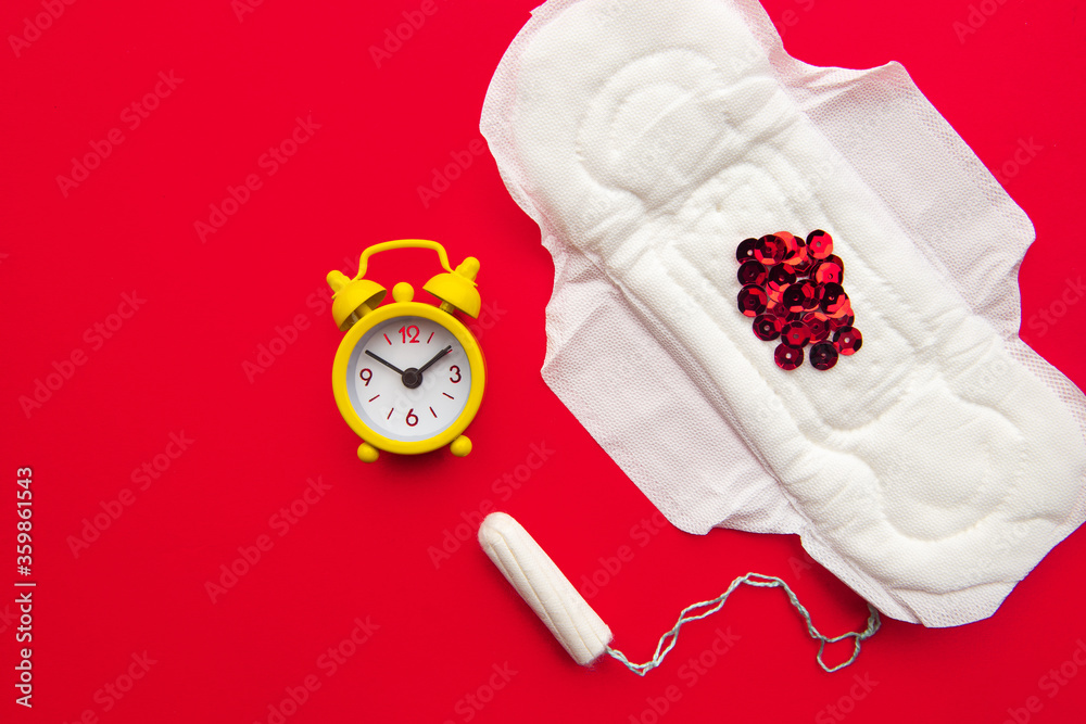 Menstrual pad with red glitter and yellow alarm clock on a red background. Menstruation period concept. Menstrual retardation concept. Menstrual pad with red glitter and yellow alarm clock on a red background. Menstruation period concept. Menstrual retardation concept.