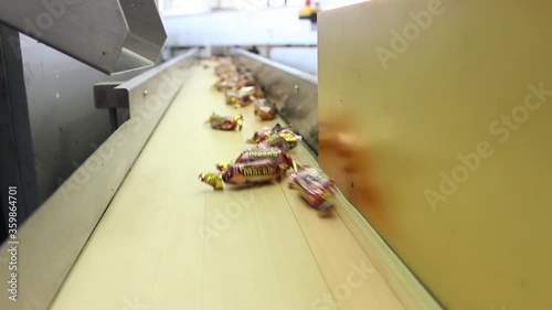 Sweets wrapped in candy wrappers fly out of a metal chute onto a conveyor belt. The video was shot in close-up.