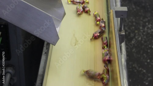 Sweets wrapped in candy wrappers fly out of a metal chute onto a conveyor belt.