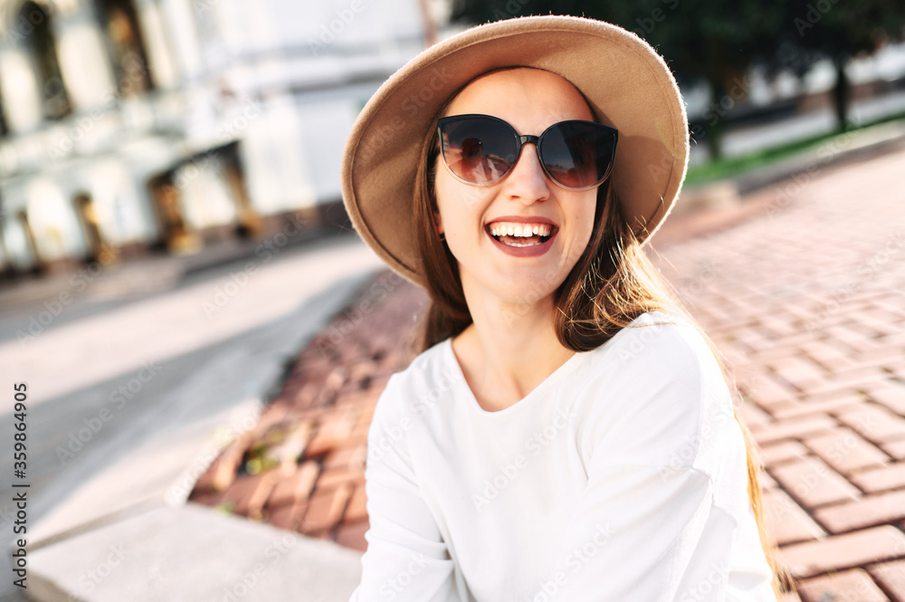 Cheerful attractive woman with sunglasses and hat is looking at camera and laughing happily outdoors. Close-up portrait in natural sunlight
