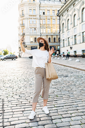 A stylish girl in mask is taking selfie on the phone or talking via video call outdoors. Young woman in mask is waving into smartphone. Full length, vertical shot