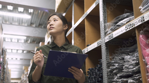 low to high portrait of warehouse worker woman looking up tall shelves doing inventory control in stockroom. female staff in uniform hold clipboard writing on paper while stock taking in storehouse
