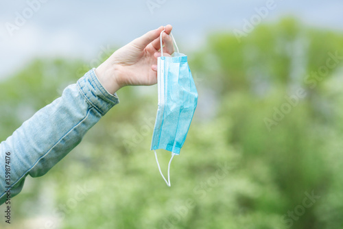 Woman takes off medical protective mask and holds it in hand on green park background. Quarantine is over. Freedom is the new life of the future
