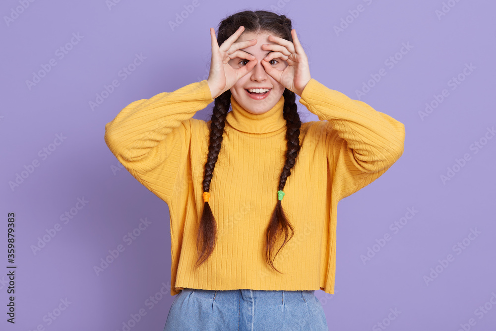 Fototapeta premium Excited brunette female with two pigtails standing with happy expression and covering her eyes with ok signs, making binocular with her fingers, looks at camera and laugh.
