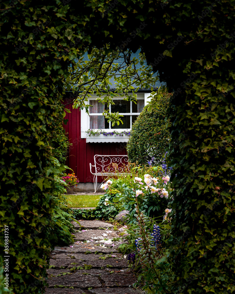 Fototapeta premium View through a an arch of ivy into a mystic garden with flowers, a bench and a red facade with white window in summer time