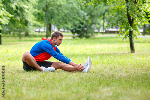 Man stretches outdoor in summer time. Sitting on a green grass.