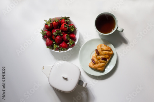 Delicious breakfast: cup of tea, tea pot, pecan bun and strawberries on white background